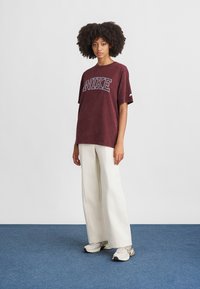 Bordo dydžio per didelis medvilnės marškinėliai su balta "NIKE" logotipu, derinami su baltais plačiais kelnais ir pilkais sportbačiais ant mėlynos grindų.