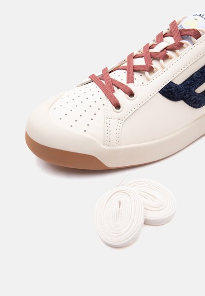 Κοντινό πλάνο μπεζ αθλητικού παπουτσιού με διάτρητο μπροστινό μέρος, καφέ κορδόνια, ναυτικό υφασμάτινο λογότυπο και δύο επιπλέον λευκά κορδόνια δίπλα σε λευκό φόντο.