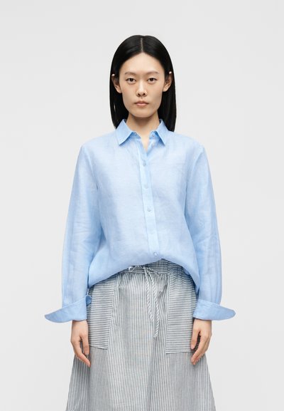 Femme aux cheveux noirs raides porte une chemise bleu clair boutonnée et une jupe grise rayée avec des poches avant sur un fond uni.