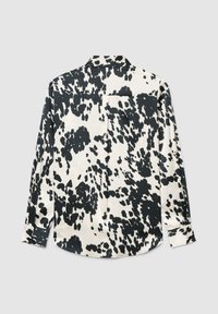 Camicia a maniche lunghe con bottoni, con motivo a macchie bianche e nere di mucca, distesa su sfondo grigio chiaro, vista dalla parte posteriore.