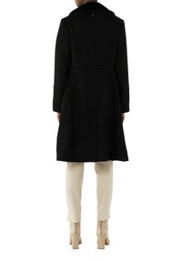 Black wool coat with a plush collar, fitted at the waist, featuring side pockets and a mid-length design, worn over cream trousers and heeled boots.
