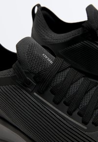 Nahaufnahme von schwarzen, atmungsaktiven Strick-Sneakern mit dicken Schnürsenkeln und strukturierten Seitenpaneelen, ausgestattet mit einer Zuglasche an der Ferse.