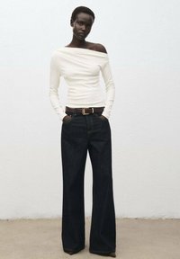 Witte off-the-shoulder top met lange mouwen, gecombineerd met high-waisted donkerblauwe wide-leg jeans. Bevat een bruine riem met een gouden gesp.