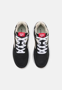 Black mesh and suede sneakers with white laces, featuring a red label and beige accents. Cushioned sole with a textured surface.