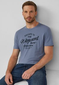 Lichtblauw katoenen t-shirt met een zwart grafisch ontwerp en tekst. Ronde hals, korte mouwen, aansluitend model. Draagt bij donkere spijkerbroek.