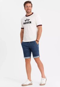 Camiseta blanca de manga corta con ribete burdeos y texto "RÊVE PARISIEN", combinada con pantalones cortos de mezclilla azul y zapatillas blancas con suelas beige.