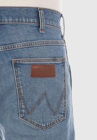 Los jeans de mezclilla azul presentan un parche de cuero con un logo en relieve, costuras en contraste y un diseño de zigzag distintivo en el bolsillo trasero.
