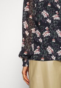 Durchsichtige schwarze Bluse mit floralen rosa und grünen Mustern, gerafften elastischen Bündchen, leicht lockerer Schnitt, getragen über einem beigen Rock.