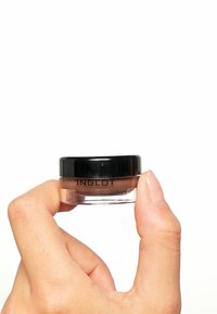 Klar glassbeholdning med brun krem og svart lokk, som viser "INGLOT"-logoen. Holdt mellom fingrene mot en hvit bakgrunn.