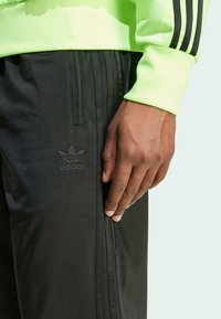 Černé sportovní kalhoty s texturovanou tkaninou, se třemi černými pruhy po stranách a embosovaným logo Adidas na stehně.