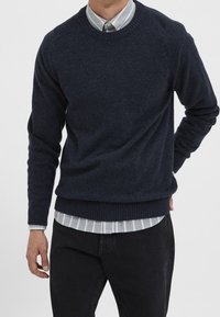 Mörk marinblå ulltröja med ribbade muddar och nederkant, designad med rund halsringning, över en ljusgrå och vit randig skjorta, bärs med svarta byxor.