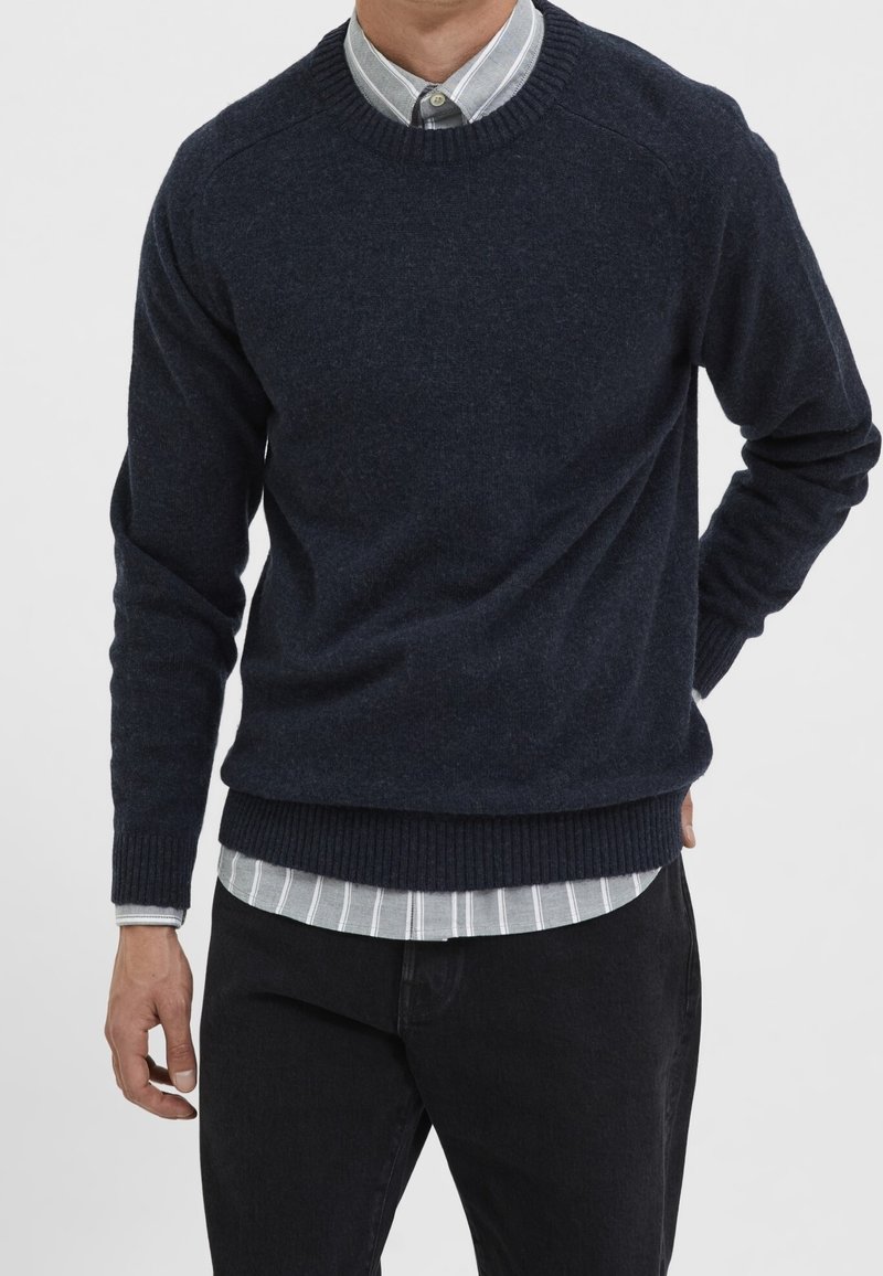 Mörk marinblå ulltröja med ribbade muddar och nederkant, designad med rund halsringning, över en ljusgrå och vit randig skjorta, bärs med svarta byxor.