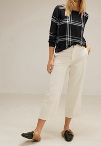 Blusa nera a maniche lunghe con motivo a quadri bianchi; pantaloni cropped color crema; mocassini neri morbidi con dettagli in pelliccia. Texture liscia e piatta.