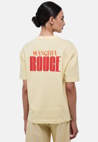 T-shirt en coton jaune avec le texte audacieux en rouge et orange "SANGRIA ROUGE" dans le dos. Manches courtes, coupe décontractée, texture douce.