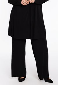 Zwart geribbeld tuniek met een vloeiende stijl gecombineerd met zwarte wijde broek. Beide stukken hebben een gestructureerde stof en subtiele beweging.