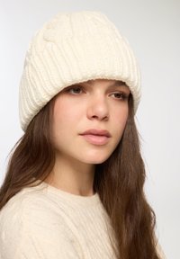 Jasno beżowa, dziana czapka z prążkowym splotem i wzorem w warkocze. Noszona swobodnie, współgrająca z swetrem w podobnym odcieniu.