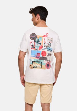 Hombre vistiendo una camiseta blanca con gráficos coloridos de playa y texto, combinado con pantalones cortos beige, visto desde atrás.