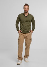 Olivgrüner Pullover mit Reißverschluss am Kragen, kombiniert mit beigefarbenen Kordhosen im Cargo-Stil mit mehreren Taschen und weißen Sneakers.