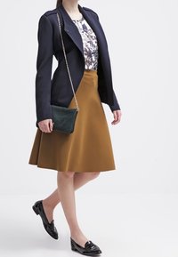 Blazer blu navy sopra una maglietta floreale, gonna A-line marrone, mocassini neri e una borsa verde con tracolla a catena. Tessuti lisci in evidenza.