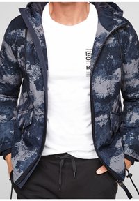 Marinblå pufferjacka med grå abstrakta mönster, dragkedjestängning, två framfickor och huva. Vit t-shirt under. Mörka byxor synliga.