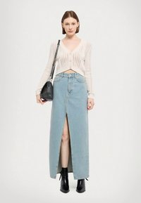 Donna che indossa un cardigan bianco corto, una gonna maxi in denim blu chiaro a vita alta con spacco frontale, stivali neri fino alla caviglia e porta una borsa a spalla nera.