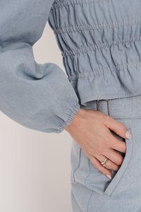 Jasnoniebieska bluzka dżinsowa z gromadzonym elastycznym detalem w talii oraz obszerne rękawy. Ręka spoczywa wewnątrz kieszeni z metalowymi elementami w kolorze srebrnym.