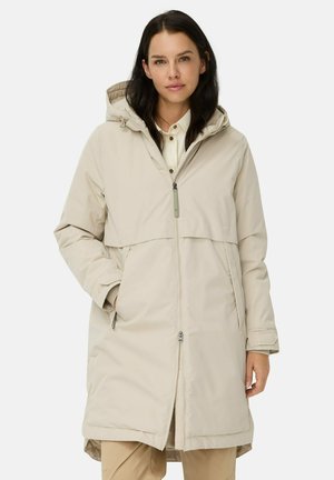 Vrouw in een beige hoodie lange jas over een witte shirt en beige broek, staand met één hand in de jaszak tegen een witte achtergrond.