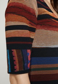 Top colorato a strisce con texture a coste. I colori principali includono rosso, nero e varie tonalità, con un accento unico sulla manica.