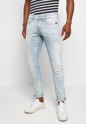 Mężczyzna ubrany w jasnoniebieskie obcisłe dżinsy podwinięte przy kostkach, białe trampki i czarno-białą koszulkę w paski, stojący na tle białej ściany.