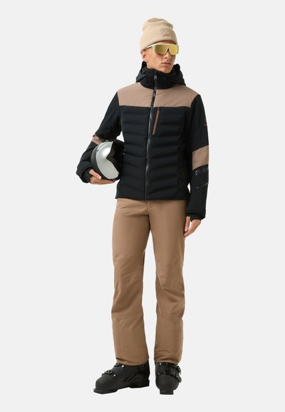 Wanderjacke in Schwarz und Beige, mit gestepptem Muster, kombiniert mit braunen Hosen. Ergänzt mit einer leichten Mütze und Sonnenbrille mit verspiegelten Gläsern. hält einen Helm.