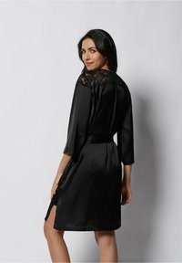 Svart satinklänning med spetsinfällning vid halsen, trekvartsärmar, skärp i midjan och en slits i sidan för rörlighet och komfort.