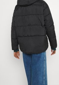 Giacca a vento nera con cappuccio, caratterizzata da pannelli trapuntati e polsini elasticizzati. Abbinata a jeans in denim blu a gamba larga.