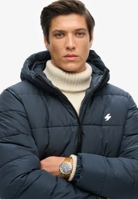 Marineblå polstret jakke med hætte og lynlås, prydet med et hvidt lynlogo. Båret over en cremefarvet turtleneck sweater.