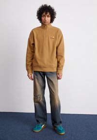 Zippered, tan sweatshirt with kangaroo pocket and embroidered logo, paired with distressed blue jeans and teal sneakers.