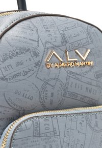 Zaino grigio con finitura strutturata, caratterizzato da un motivo a timbro. Presenta dettagli in oro e un logo con la scritta "ALV by Alviero Martini."