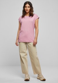 Lichtroze T-shirt met korte mouwen en een losse pasvorm, gecombineerd met beige wijde broekspijpen. De outfit wordt compleet gemaakt met sportieve schoenen in neutrale tinten.