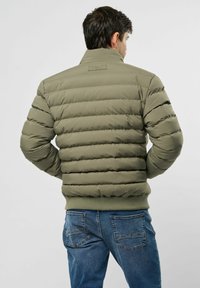 Muškarac okrenut leđima nosi maslinasto zeleni prešani puffer jaknu i plave traperice, nasuprot jednostavnoj svijetloj pozadini.
