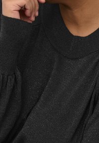 Svart pullover-tröja med ribbade muddar och krage, med en mjuk textur och subtila glittrande accenter. Synlig hand som vilar på hakan.