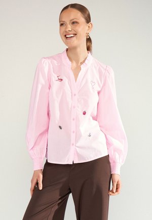 Frau lächelt und trägt eine hellrosa, geknöpfte Bluse mit Puffärmeln und kleinen Insekten- und Herzstickereien, kombiniert mit dunkelbraunen Hosen.