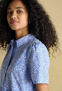 Sinine lühikeste varrukatega särgiga, millel on valge leopardimuster ja kogumisega detailid õlgadel. Sile tekstuur.