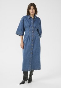 Denimist kleit, millel on nööpidest ees, lühikesed varrukad ja pikk, lõdva lõikega siluett. Omab vöökohtade rõhumist ja kontrastseid õmbluse detaile.
