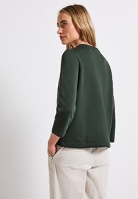 Maglione di cotone verde scuro con scollatura rotonda e maniche a tre quarti, abbinato a pantaloni beige chiaro. Texture semplice e liscia.