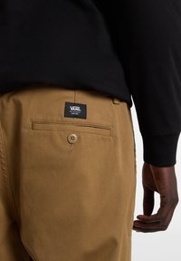 Beżowe materiałowe spodnie z tylną kieszenią z zapięciem na guzik oraz metką z logo Vans. Czarna koszula widoczna powyżej linii talii.