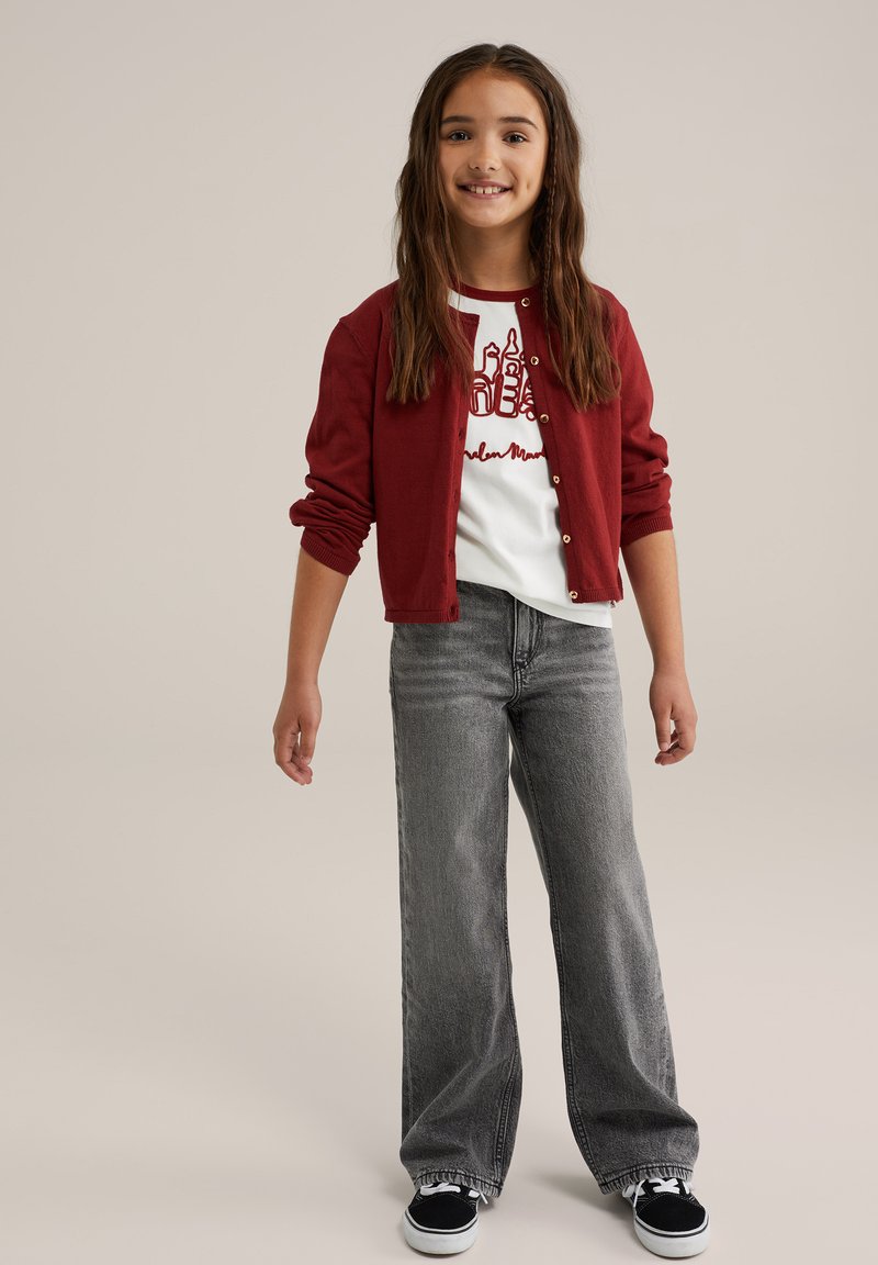 Tyttö seisoo pitkät ruskeat hiukset, päällään punainen neuletakki, valkoinen graafinen t-paita, harmaat leveälahkeiset farkut ja mustat lenkkarit.