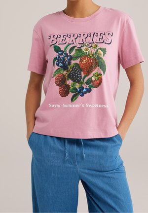 Osoba w różowej koszulce z kolorowym nadrukiem jagód oraz napisem "BERRIES Savor Summer's Sweetness", połączona z niebieskimi spodniami na sznurki.