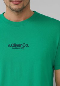 Man in een groen T-shirt met "s.Oliver Co. Established 1969" in het zwart op de borst gedrukt.