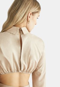 Top corto beige chiaro con colletto, caratterizzato da un'apertura a chiave sulla schiena, orlo arricciato e una texture di tessuto liscia e leggermente lucida.
