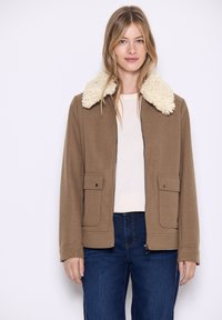 Giacca di pile marrone con un colletto soffice color crema, dotata di due tasche frontali e chiusura con zip, indossata sopra una maglietta chiara e jeans.