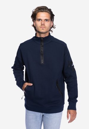 Man met golvend bruin haar draagt een marineblauwe pullover met ritsen in zwart en lichtblauwe spijkerbroek, staand tegen een effen achtergrond.