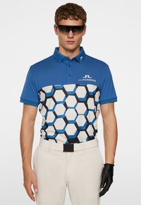 Blå polo med ett geometriskt hexagonalt mönster i svart och ljusblått, kortärmad, och en J.Lindeberg-logotyp. Bärs med beige byxor och svarta handskar.